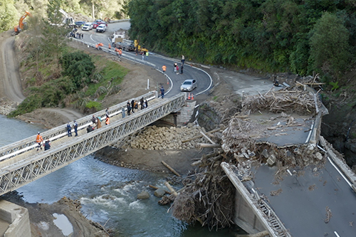 A destroyed bridge due to a landslide, with debris scattered around the area. Next to the destroyed bridge is a newly built bailey bridge with construction crew and people inspecting it.