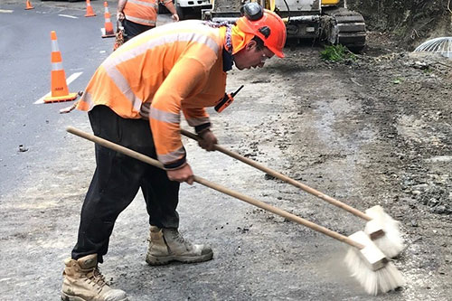 A man in an orange shirt and safety vest sweeping the road to maintain cleanliness and ensure safety.