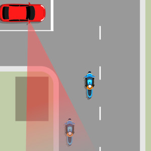 A red car at an intersection with a shaded red area showing the car's blind spot. 2 blue motorcycles are traveling on the intersection road. one is in the blind spot and one can be seen..