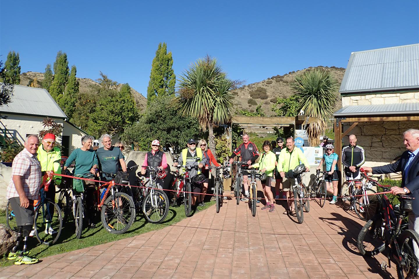Group of cyclists standing with their bikes with a red ribbon strung across the brick path at the opening of the Hurunui Heartland Ride.