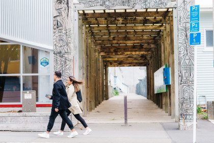 Man and woman walking along a street pass a laneway with wooden boards.