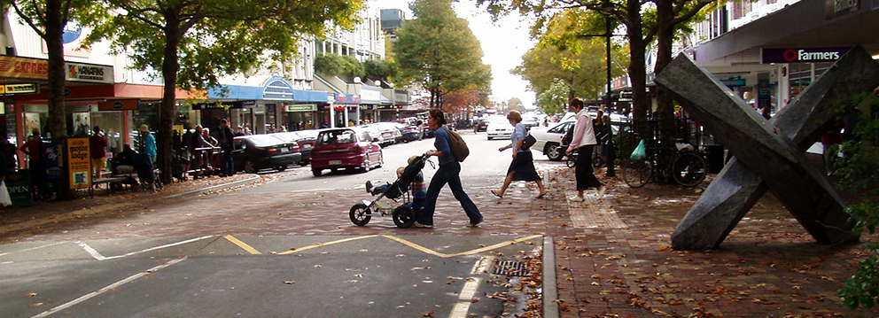 Woman pushing a pram over an elevated pedestrian crossing in a leafy high street during Autumn.