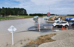 White car and milk tanker stopped at intersection stop sign as another milk tanker drives through the flooded highway.