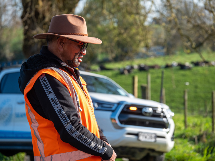 A man wearing an orange vest and hat stands before a white truck.