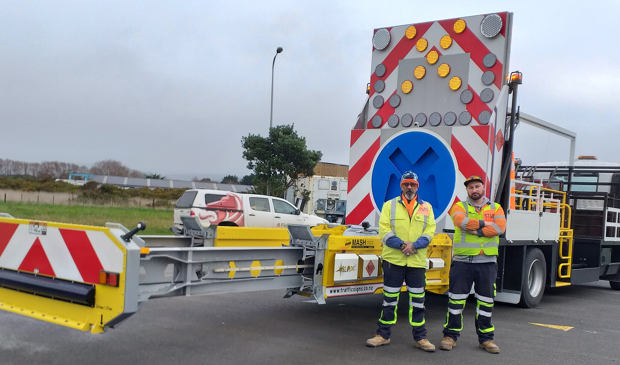 Truck with trailer on the back with illuminated traffic management signals and two men standing with yellow and orange high-vis gear on.