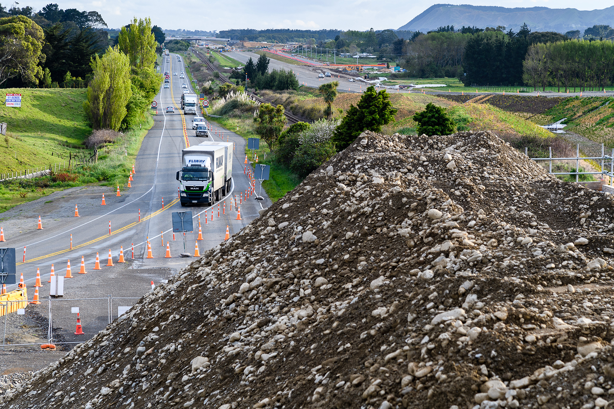 Large pile of dirt in the foreground with traffic separated by road cones.