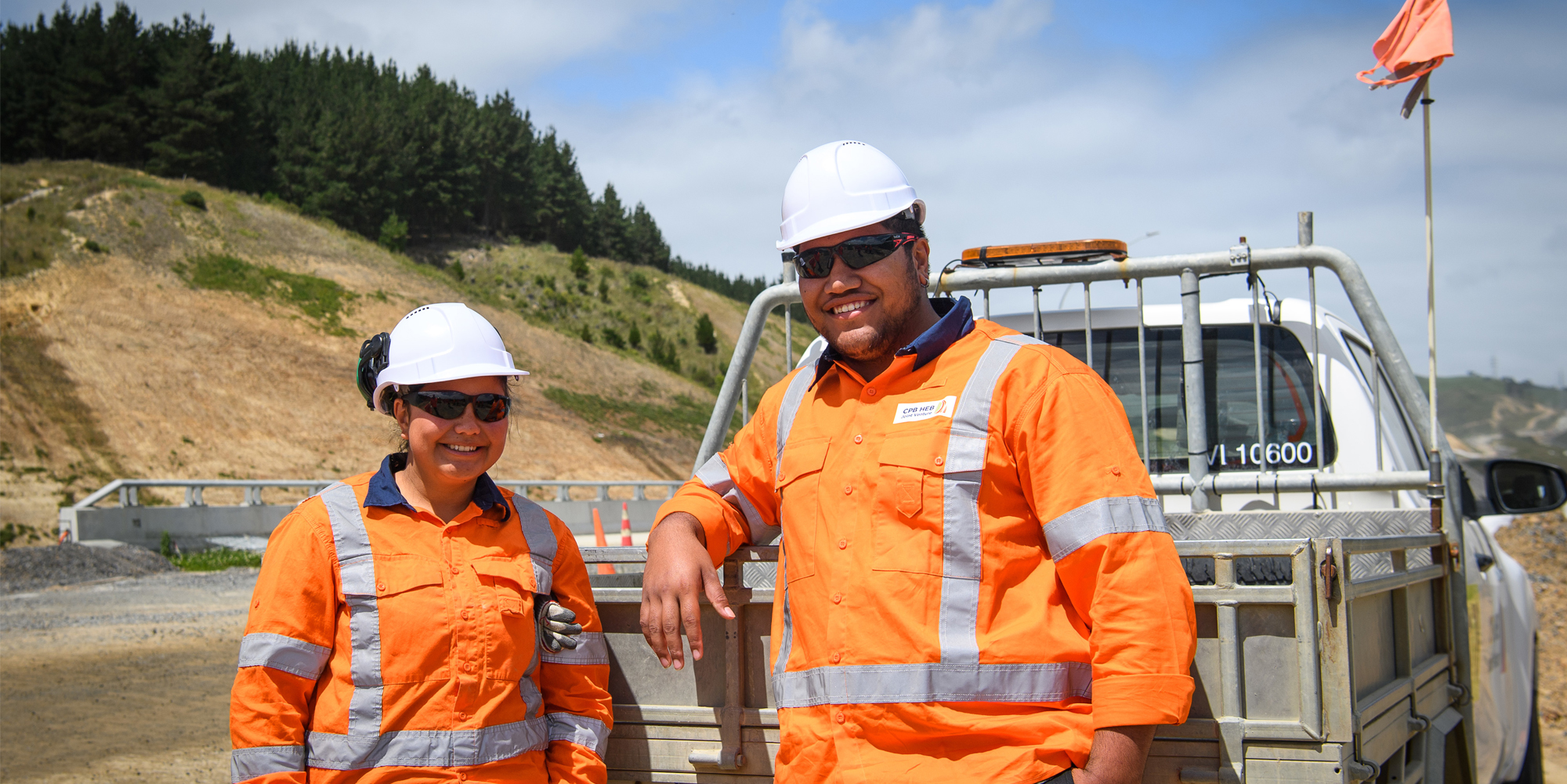 Two construction workers smiling and standing in front of a truck on site.