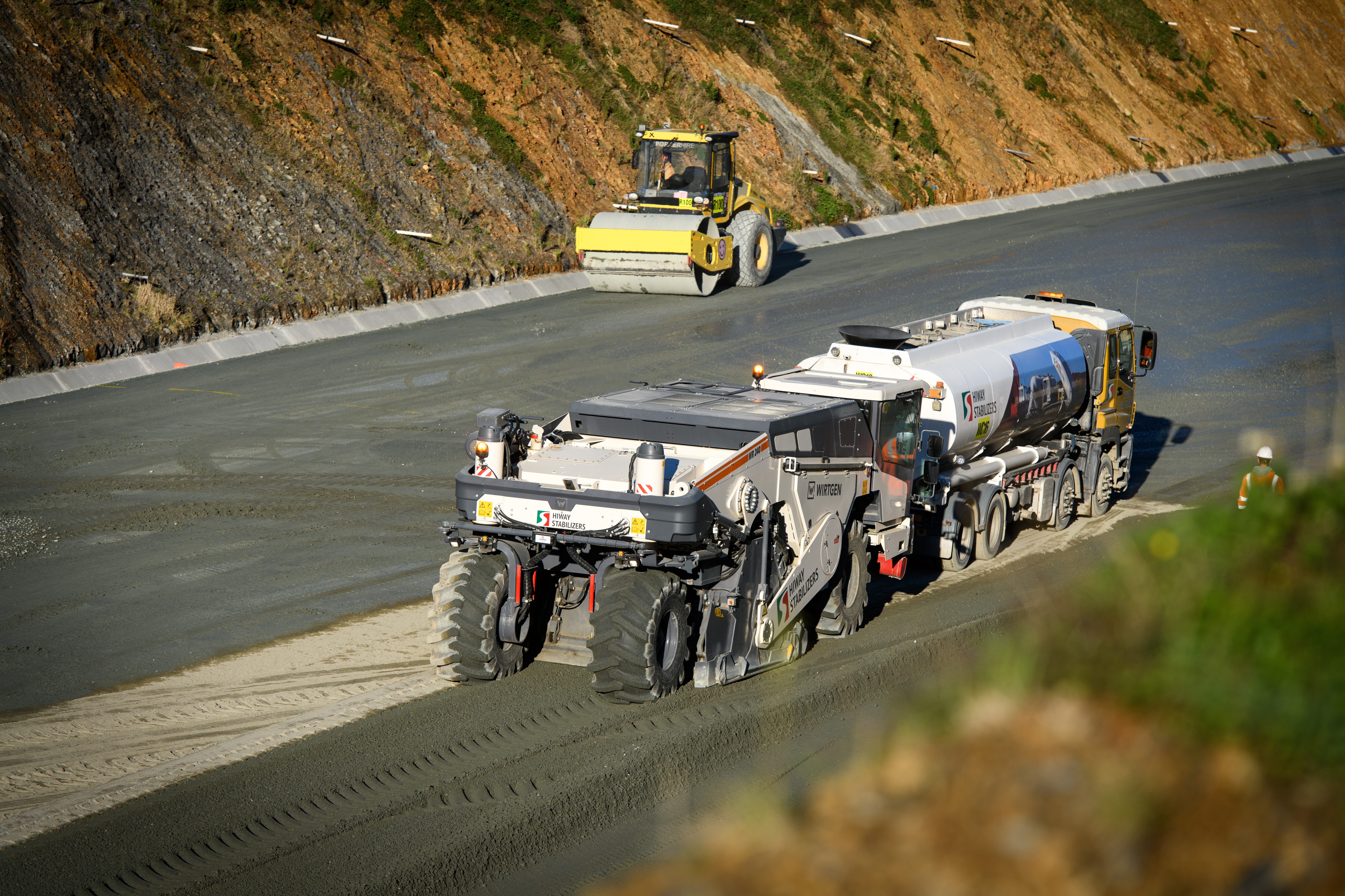 Stabiliser following water cart moves over surface mixing cement with aggregate