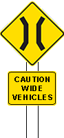 Two permanent warning signs combined on one post, the top one showing a narrow bridge icon and the other says caution wide vehicles. Both signs have a yellow background