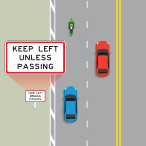 A blue car, a red car and a motorcycle are traveling along a 4 laned road with double yellow lines in the centre. A sign on the grass verge is enlarged so you can read the sign. The sign says: Keep left unless passing.