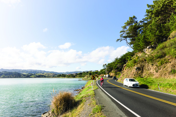 An asphalt road running alongside a body of water. A cyclist is riding away from the camera and a white car is travelling toward the camera.