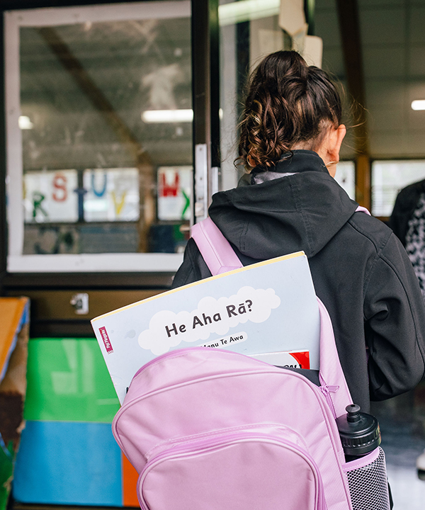 A child walking into a classroom, with a te reo Maori book in her school bag