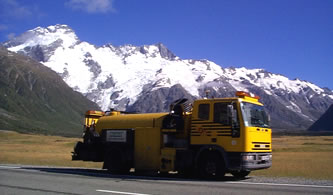 A yellow SCRIM truck is stationary on the shoulder of a state highway road. It's a sunny day with some visible snow mountains in the background.