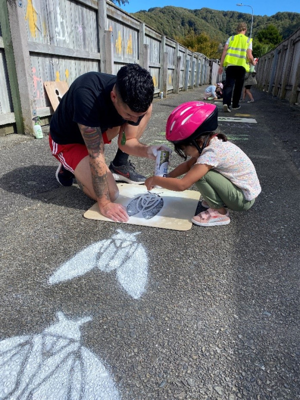 A person and a child stencilling a cicada design on the ground