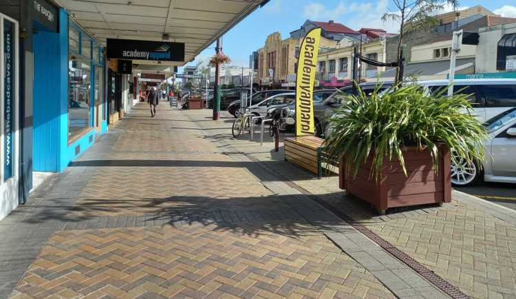 A wide footpath along a shopping street. A large, wooden planter box and bench are at the roadside edge of the footpath.