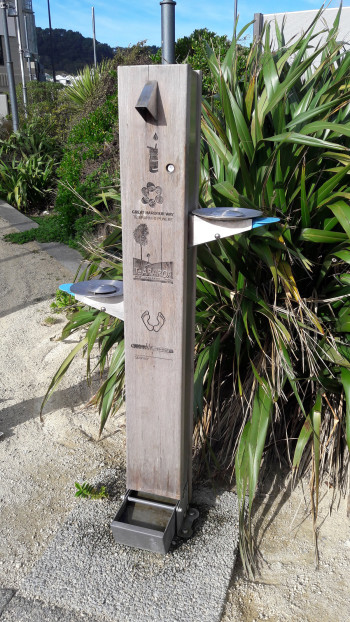 A wood finished water fountain at the edge of a path with different heights for users.