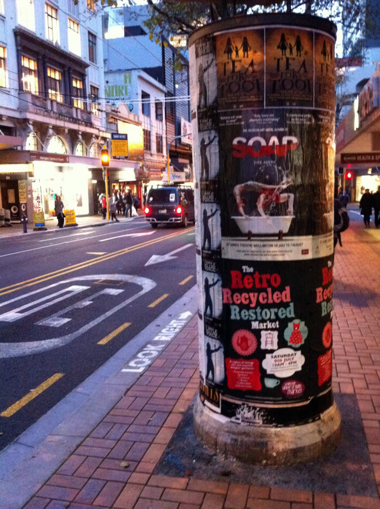An advertising pillar, covered in posters, near the edge of the footpath.