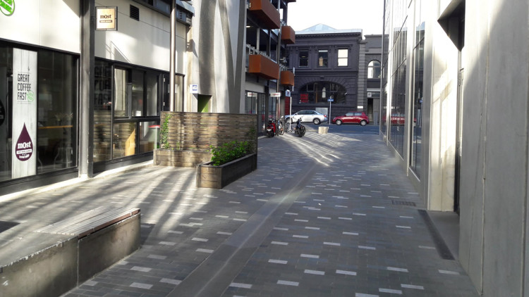 Outdoor dining space and vehicle access in Christchurch laneway, with a sidewalk and bench.