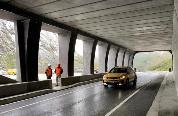Yellow car driving on freshly sealed road inside the entrance to the tunnel with two workers in high visability jackets and helmets looking on.