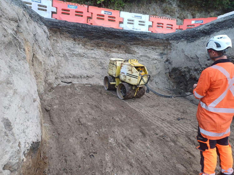A man in an orange shirt and pants operates a remote-controlled mini padfoot trench roller on a construction site.