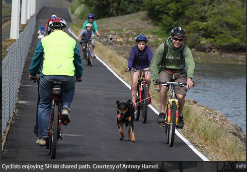 Smiling cyclists pass each other on the SH88 shared path between Dunedin and St Leonards. One cyclist has a dog.
