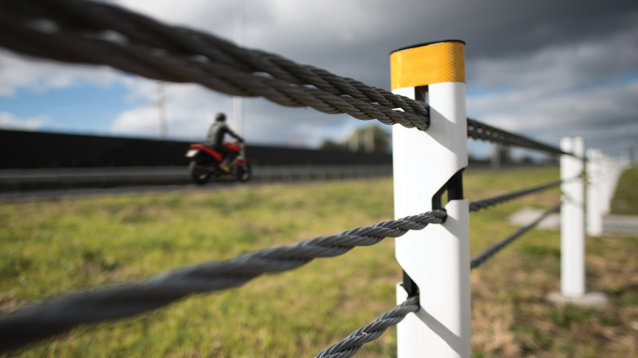 Looking through wire rope barrier at motorcyclist riding on rural road.