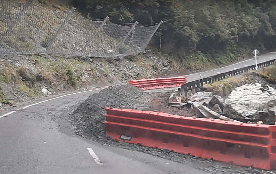 Road reduced to one lane with nets on the upper sloped bank on the left to stop rocks falling on the road and a section of the collapsed road on the right.