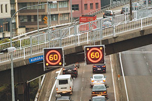 2 alnes of cars travelling on the motorway. A bridge goes over the motorway. 2 lighted speed signs are attached to the bridge and show the speed the vehicles are allowed to travel.
